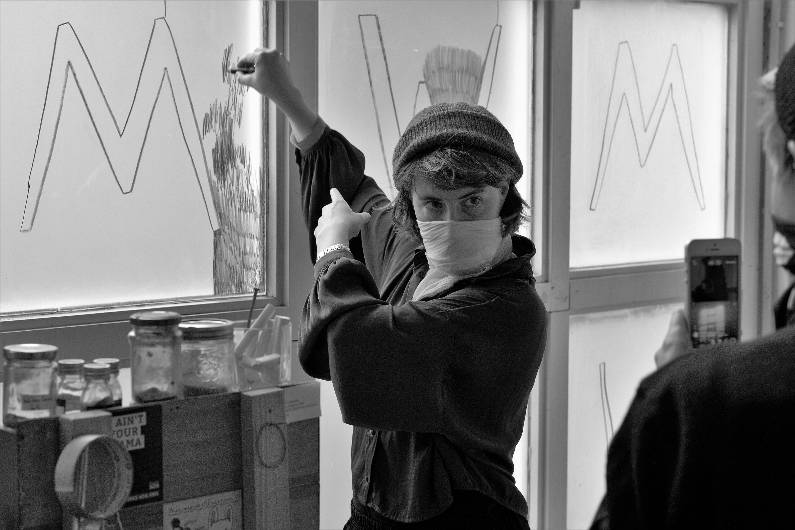 picture shows Marie aka Luis performing blù at their graduation show M. Luis is holding a blù wax pen with their right hand blind drawing lines on window glass, to frame the left out letter M in the middle of the window while facing to their left side where their performance fighter Izzy is standing, watching while holding a phone which is live streaming this action.

picture taken by Martin Hoffmann 